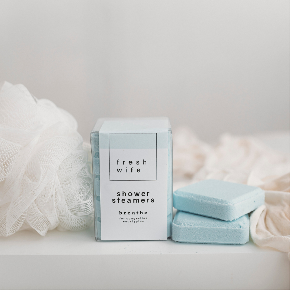 A boxed shower steamers product labeled 'fresh wife' stands on a white surface with two pale blue bath blocks beside it. Soft white and beige fabric props and a white mesh loofah are arranged around it against a light neutral background.