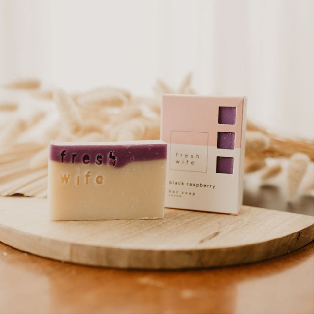 A handmade bar of soap in a cream and purple colorway, embossed with the brand name 'fresh wife', stands next to its light pink packaging box on a round wooden board.
